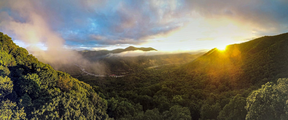 A mountainous landscape with dense forest, a winding road, and a sunrise or sunset with clouds.