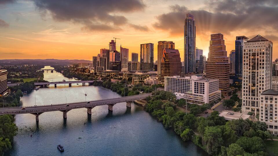 A cityscape with a river, a bridge, and high-rise buildings during sunset.
