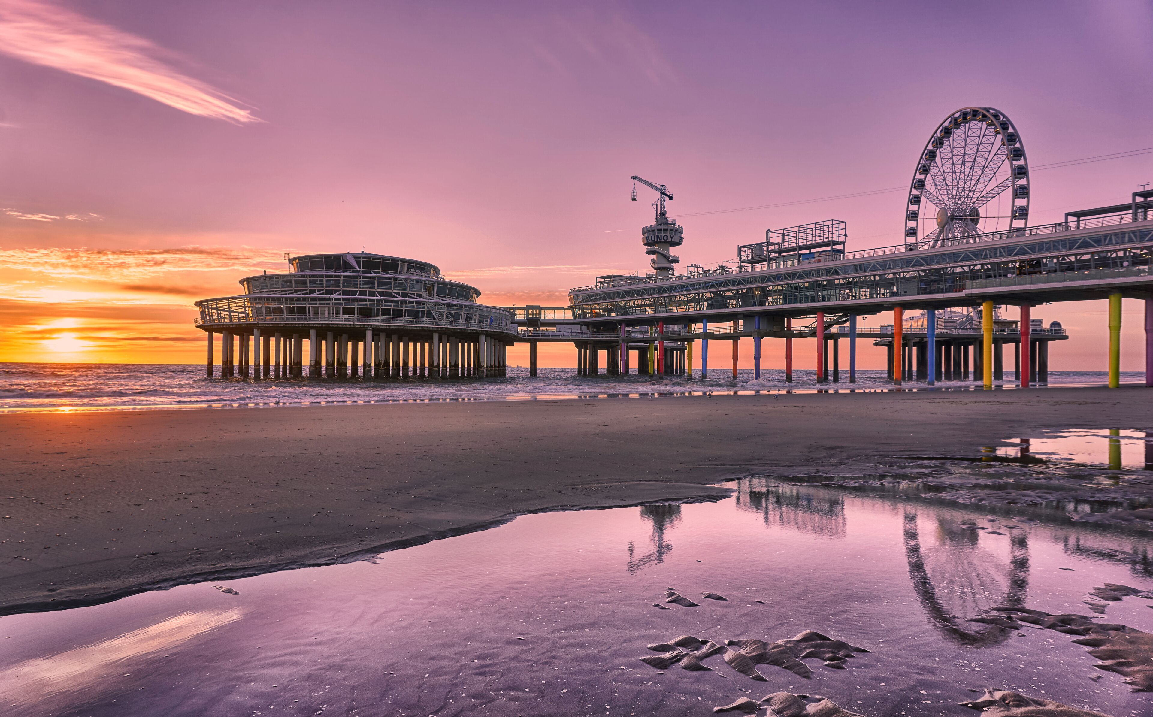 A pier with a Ferris wheel and a circular building at sunset.