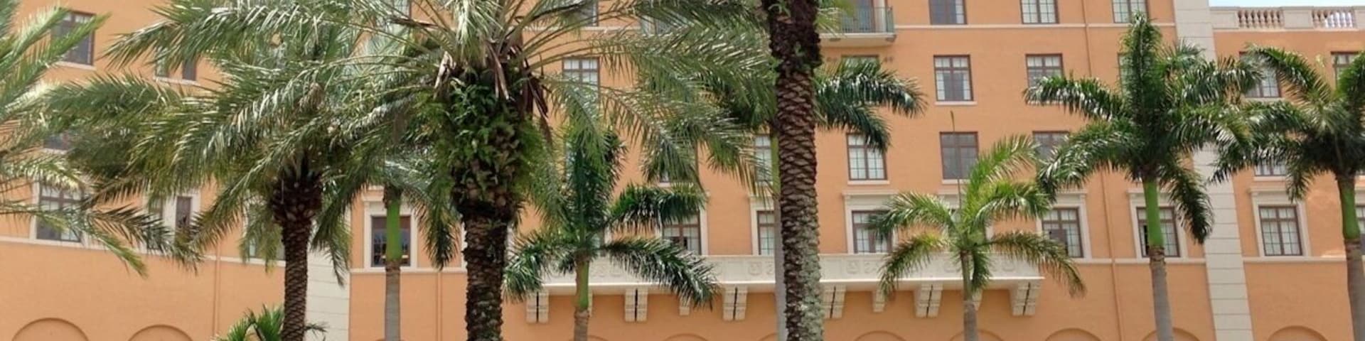 Un immeuble à plusieurs étages, doté d’une façade de couleur pêche, entouré de palmiers et d’une végétation luxuriante et bien entretenue.