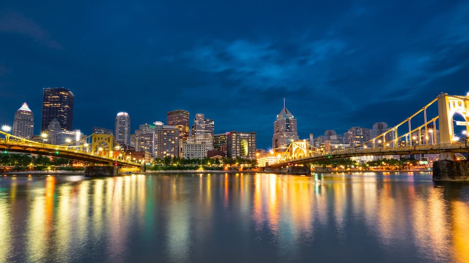 A city skyline at night with illuminated buildings and a bridge over a river.