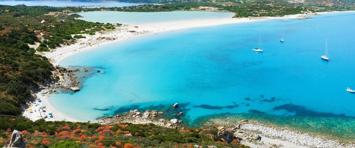 A coastal landscape with a sandy beach, clear turquoise waters, and boats anchored near the shore.