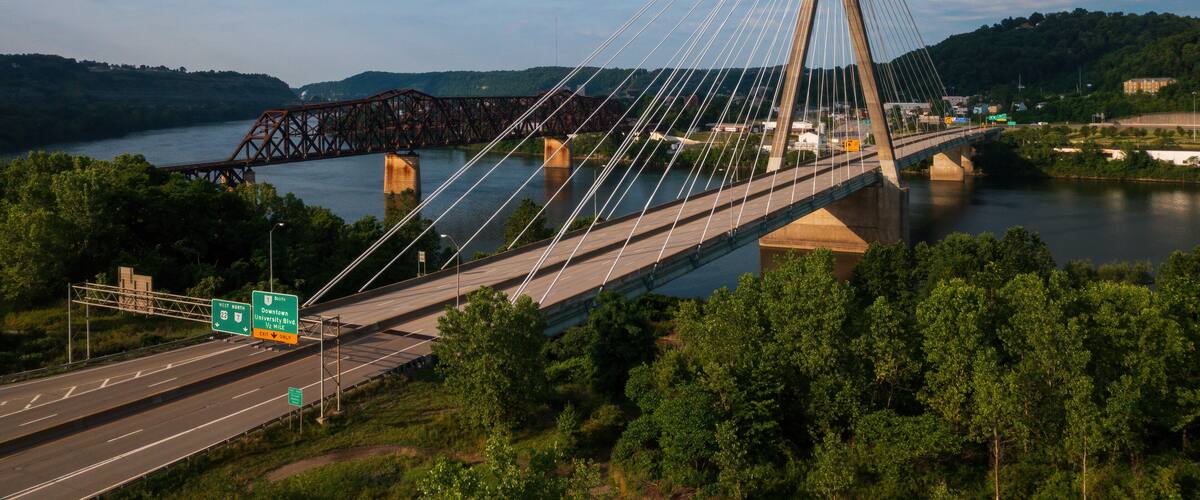 A cable-stayed bridge over a river with a forested hillside in the background.