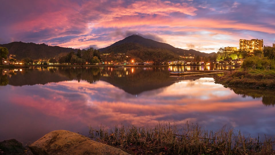 A mountain at sunset with a lake reflecting the sky, a bridge, and buildings on the right.