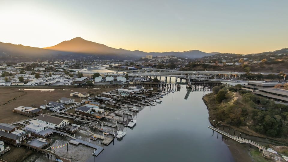 A marina with boats docked, a bridge, and a mountainous backdrop.