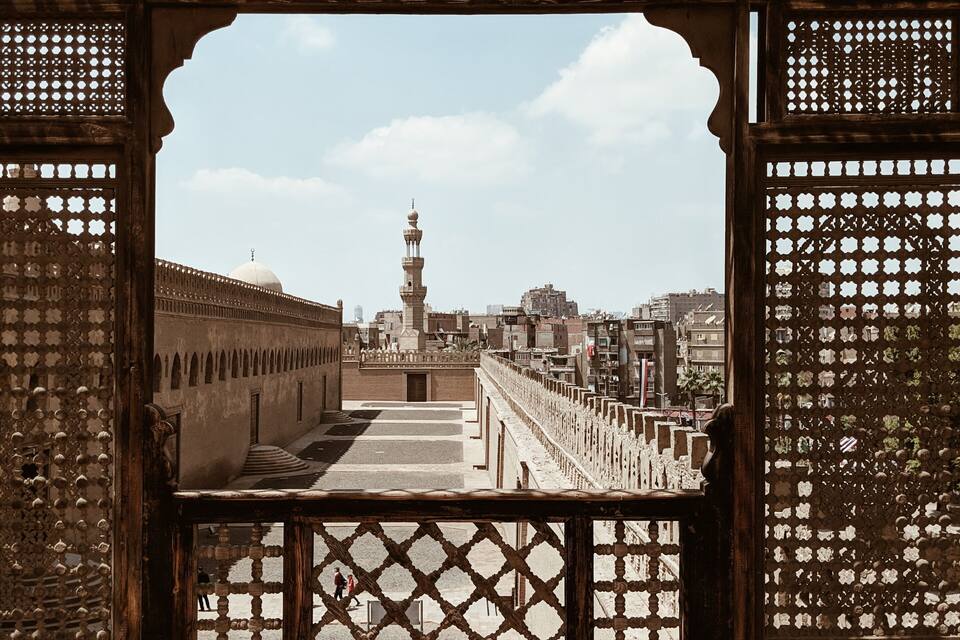A view through a wooden lattice structure with a mosque and minaret in the distance.