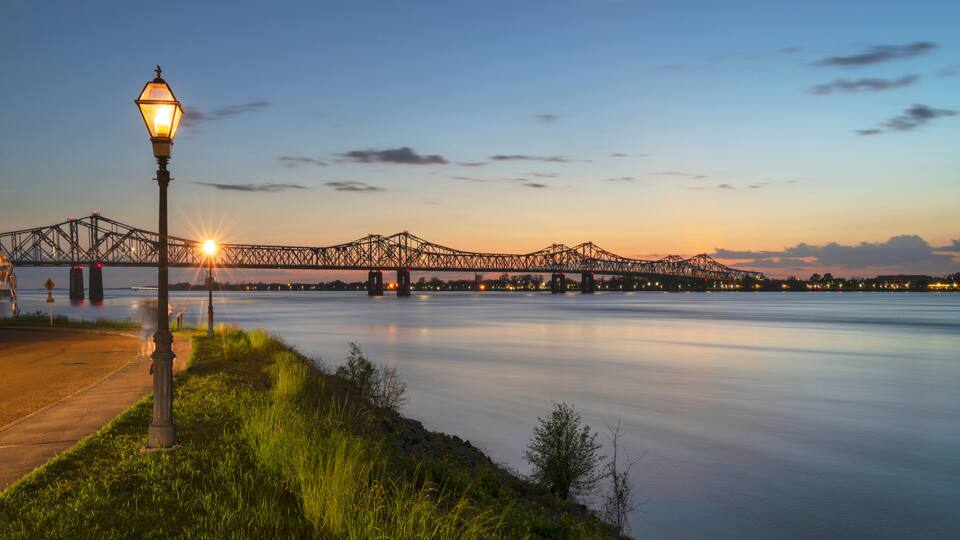 A sunset over a river with a bridge, a street lamp, and a grassy bank.
