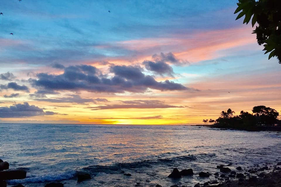 Un atardecer sobre una costa rocosa con palmeras silueteadas contra el cielo.