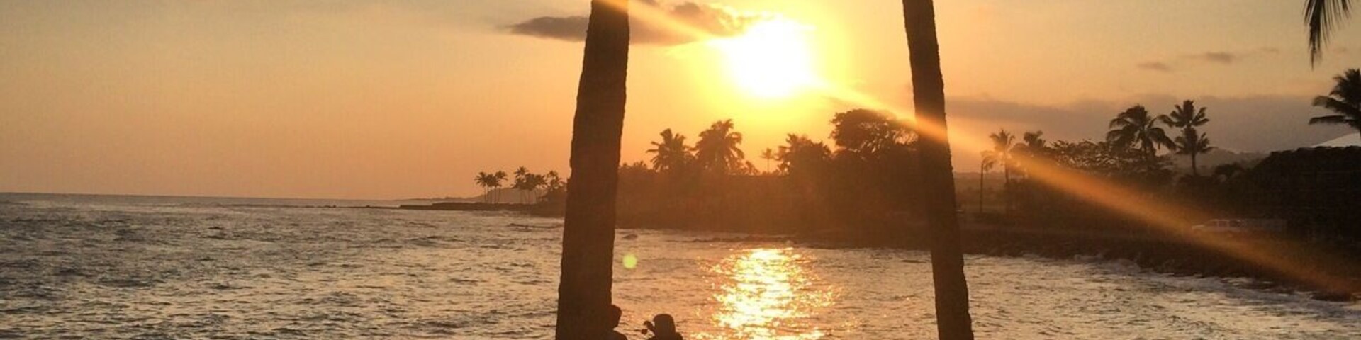 Un atardecer sobre una playa con palmeras y gente sentada en la orilla.