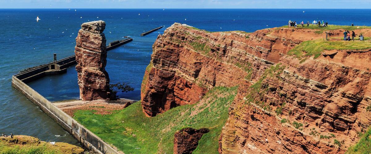 A coastal cliff with a lighthouse, a pier, and people on the cliff top.