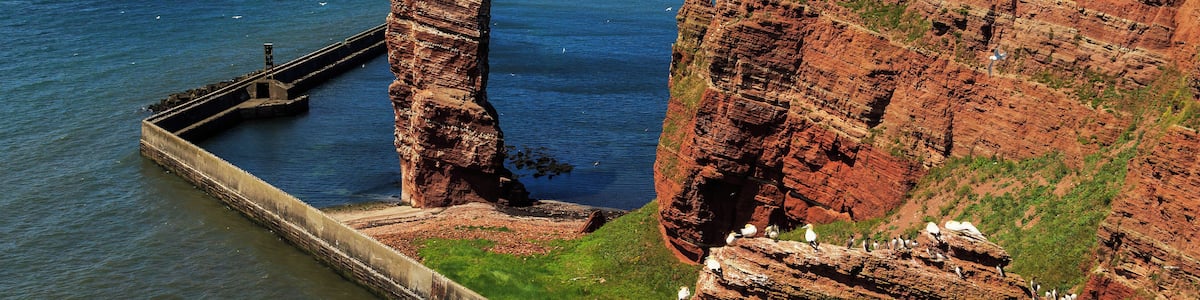 Eine Küstenlandschaft mit einer markanten Felsformation, einem Steg, der sich ins Meer erstreckt, und einem fernen Leuchtturm.