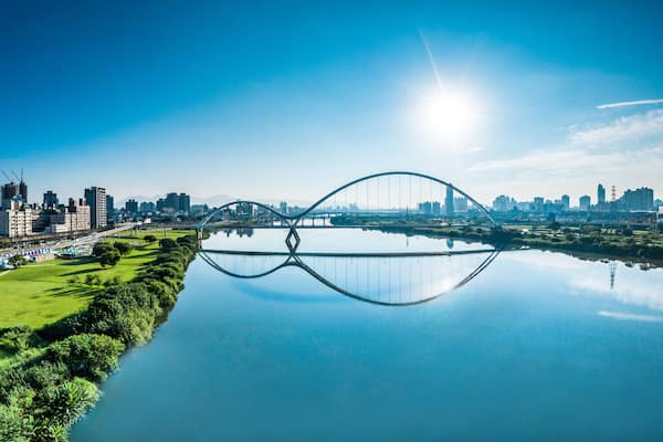 Eine Stadtansicht mit einer großen Brücke über einem ruhigen Fluss, der den Horizont spiegelt.