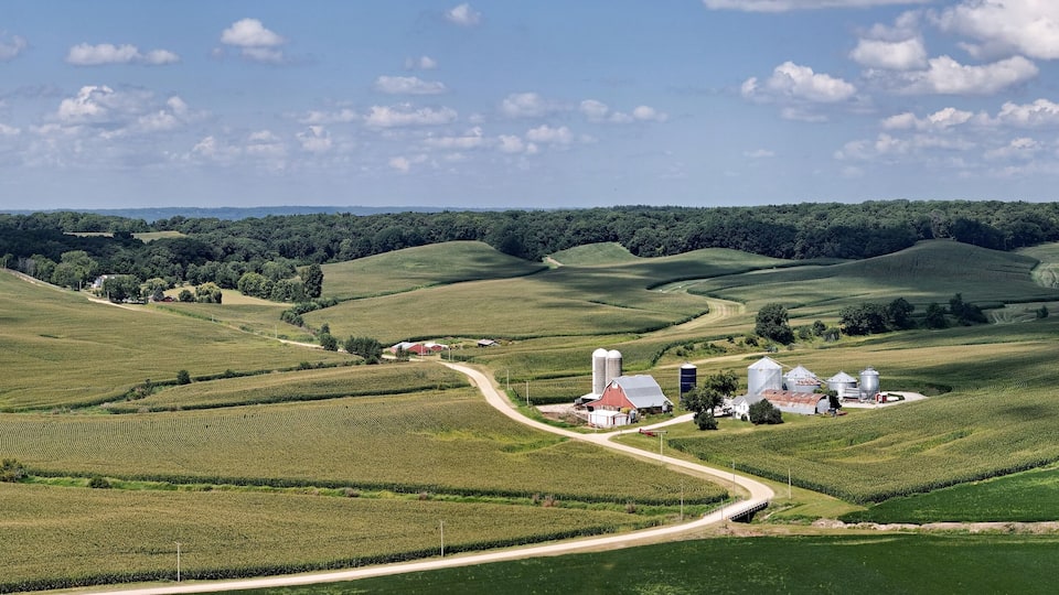 A rural landscape with rolling hills, a winding road, and a farmstead with silos.