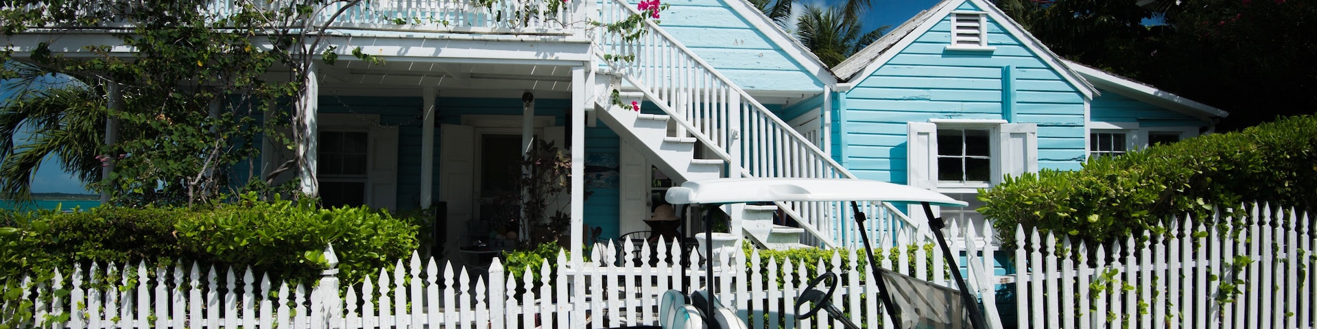 A turquoise car parked in front of a white picket fence, a blue house with a porch, and palm trees in the background.