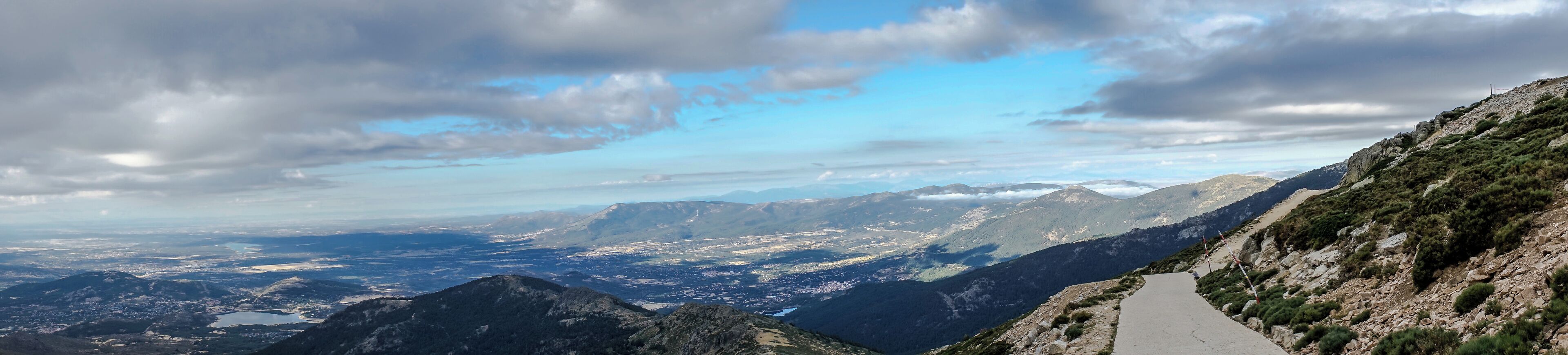 A mountain road winding through a valley with distant mountain ranges under a partly cloudy sky.