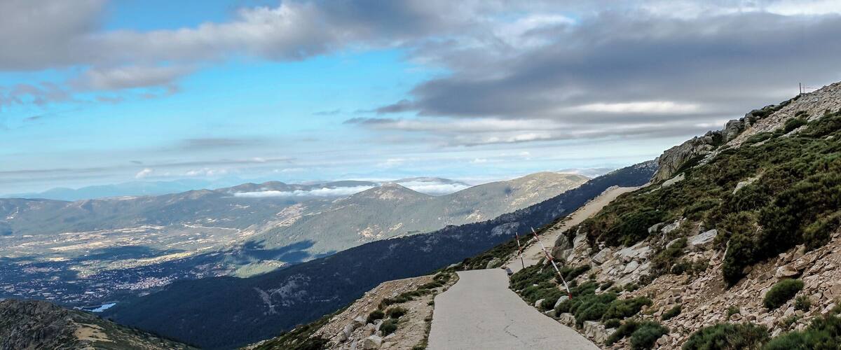 A mountain road winding through a valley with distant mountain ranges under a partly cloudy sky.