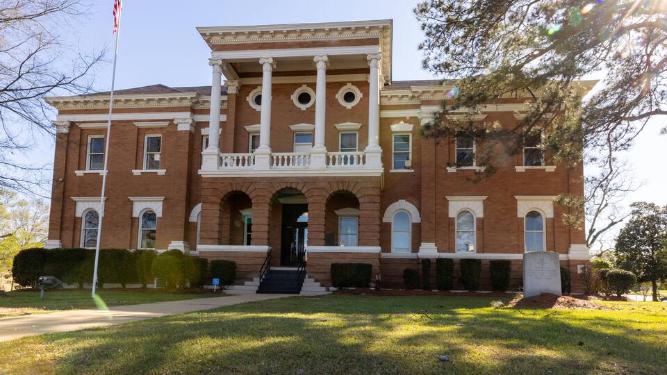 A large brick building with a central entrance, columns, and a flagpole.