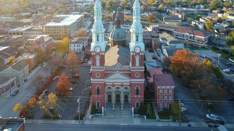 A historic church with two towers and a central dome, surrounded by autumn-colored trees and a cityscape.
