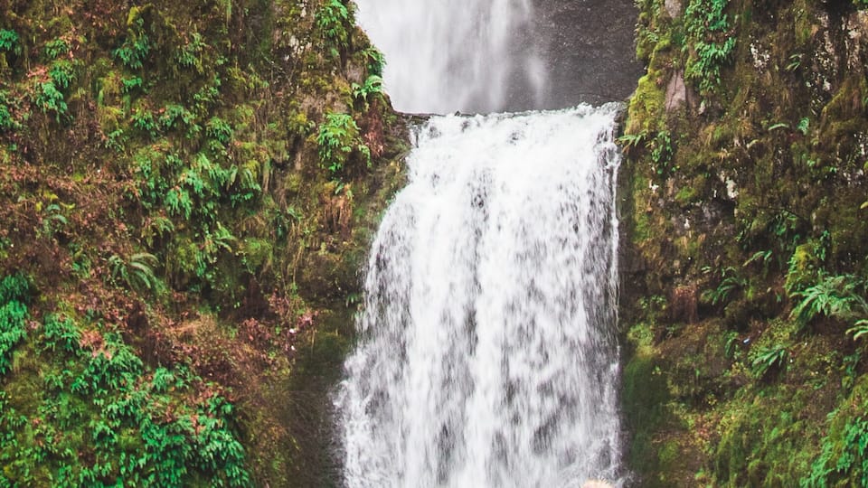 A waterfall under a bridge with two people observing.