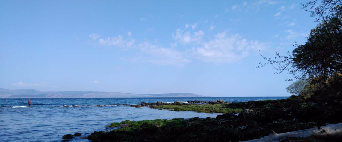 A rocky shoreline with calm sea, distant mountains, and a clear sky.
