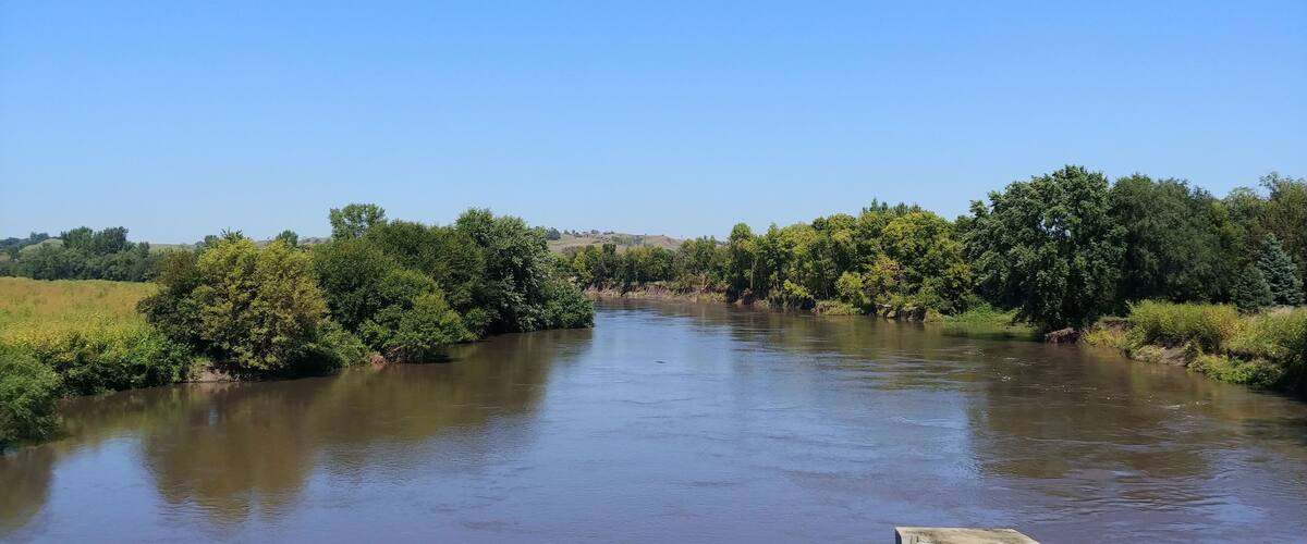 A river with dense greenery on both sides, a clear blue sky, and a concrete barrier in the foreground.