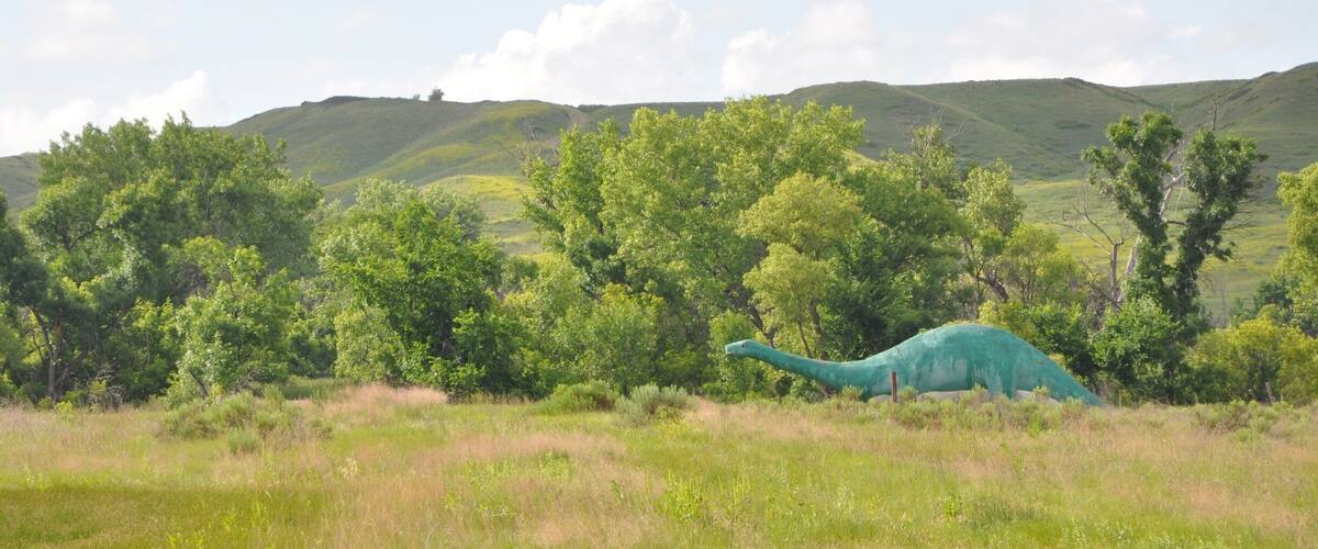 A green dinosaur sculpture in a grassy field with hills and trees in the background.