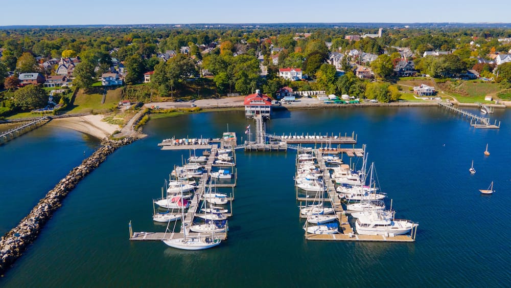 A marina with multiple boats docked, surrounded by a residential area and a clear sky.
