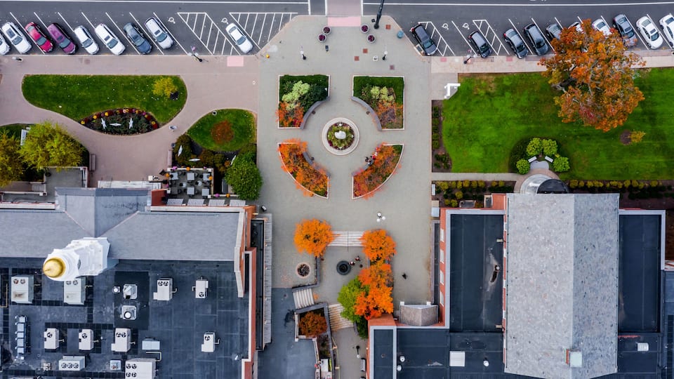 An aerial view of a parking lot with multiple rows of cars, green landscaped areas, and a central circular feature with trees.