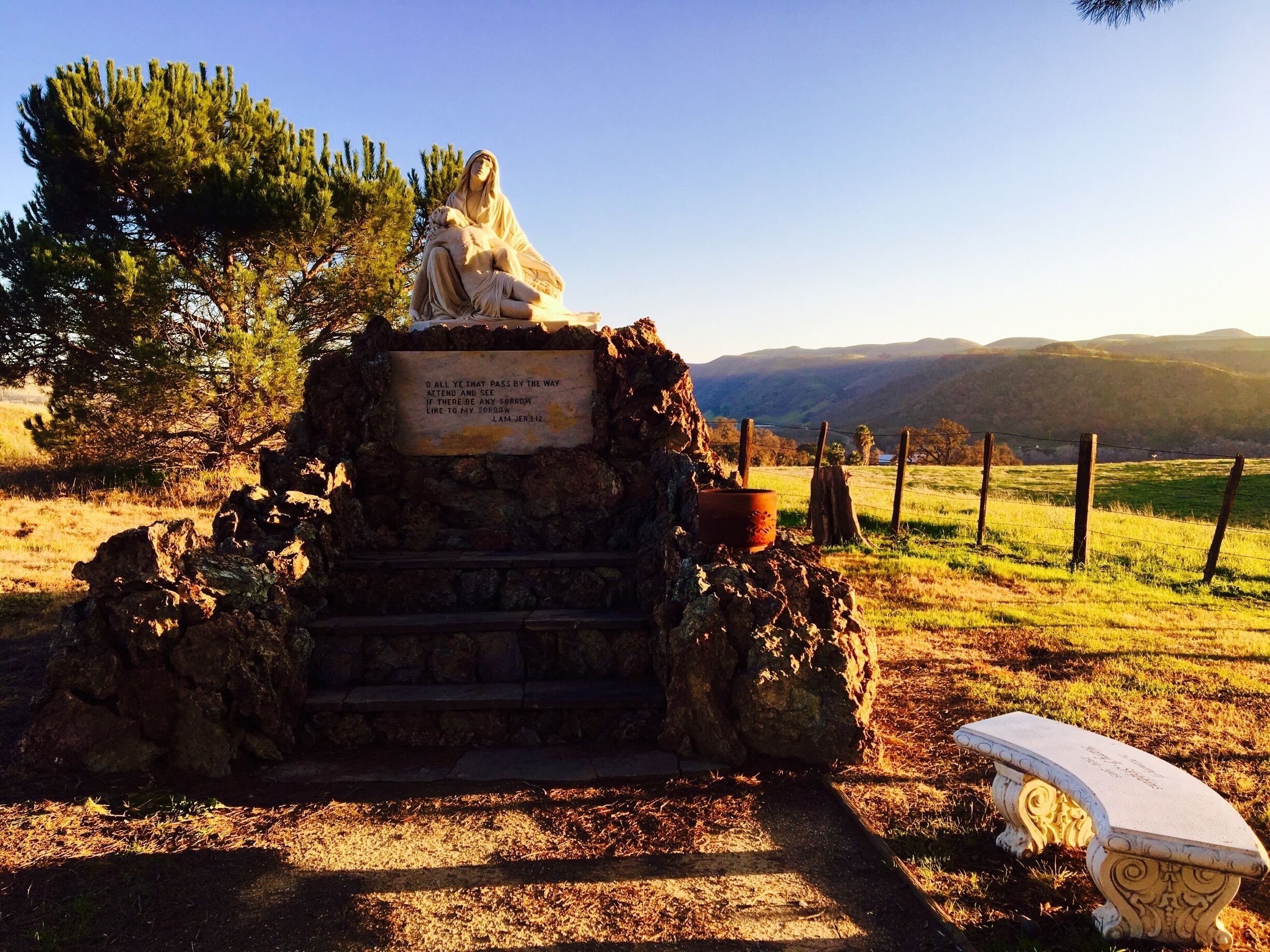 A statue of a woman holding a child on a stone pedestal with a plaque, surrounded by a rural landscape with trees and hills.