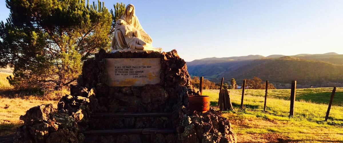 A statue of a woman holding a child on a stone pedestal with a plaque, surrounded by a rural landscape with trees and hills.