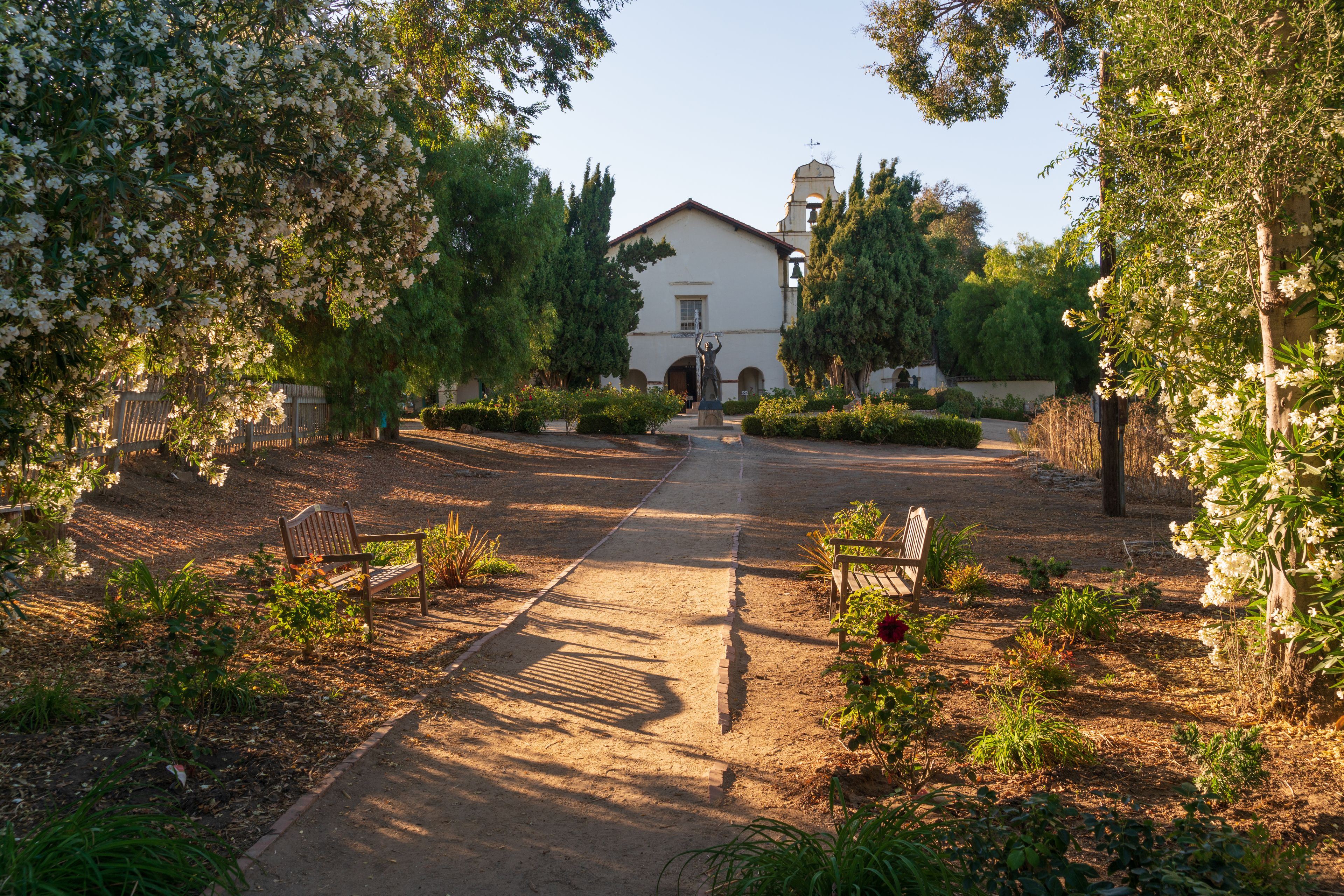 A pathway leading to a white building with a bell tower, surrounded by flowering trees and shrubs.