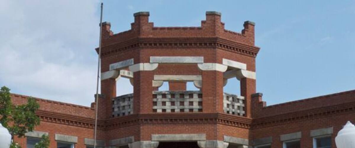 A brick building with a central tower and a balcony, surrounded by a pathway and street lamps.
