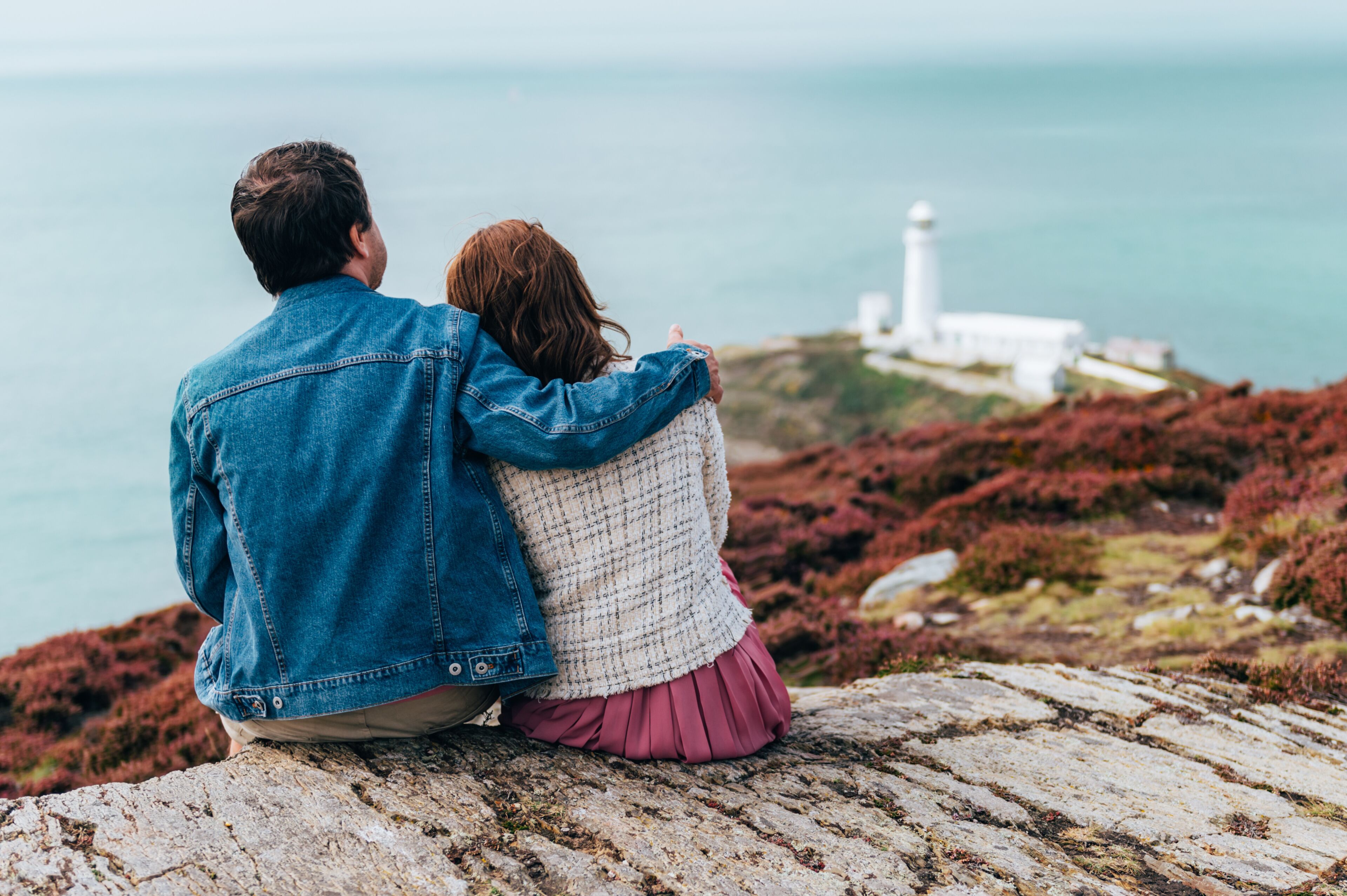 Una pareja sentada sobre un afloramiento rocoso, con vista al mar y un faro a lo lejos.