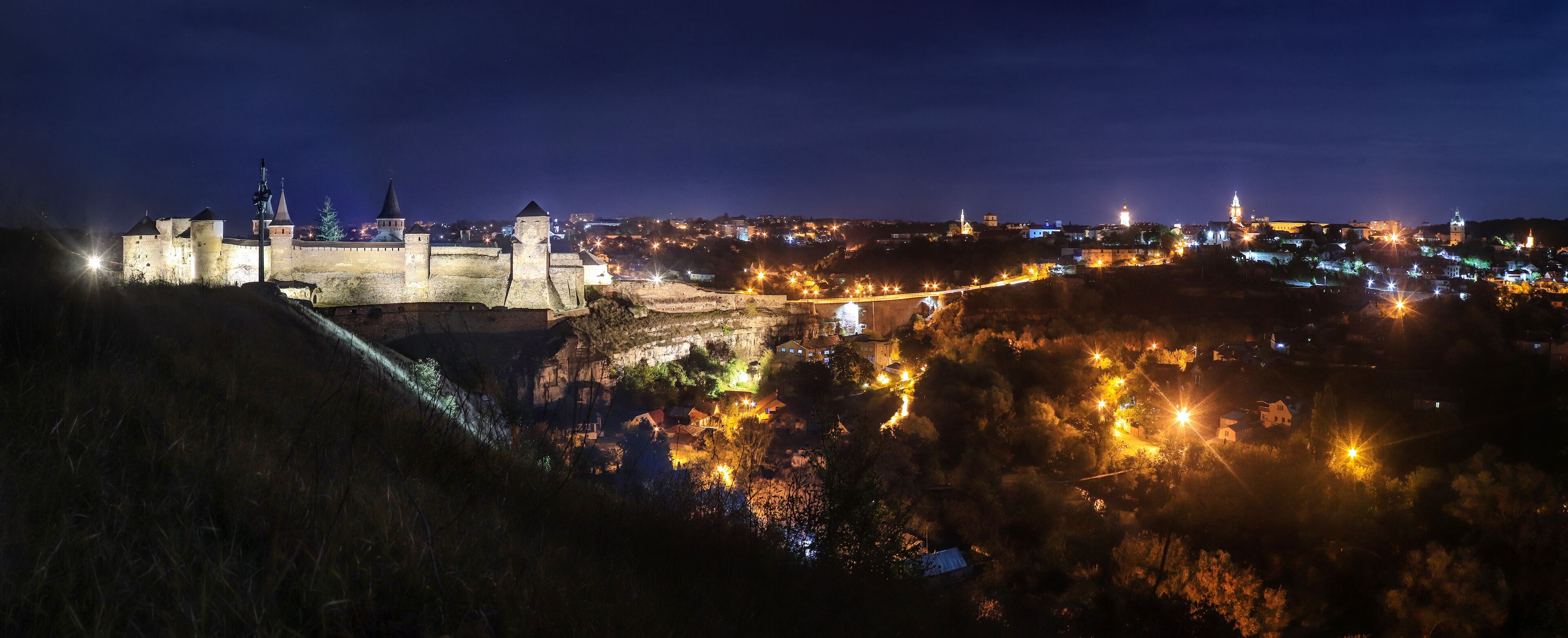A historic castle with a moat, surrounded by a town with illuminated buildings at night.