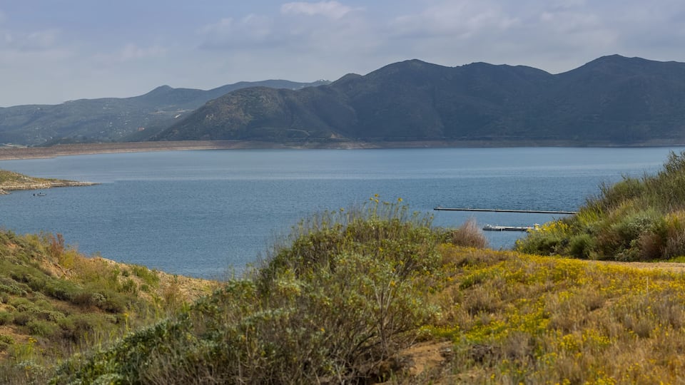 A coastal landscape with a dirt path, a calm lake, and mountains in the background.
