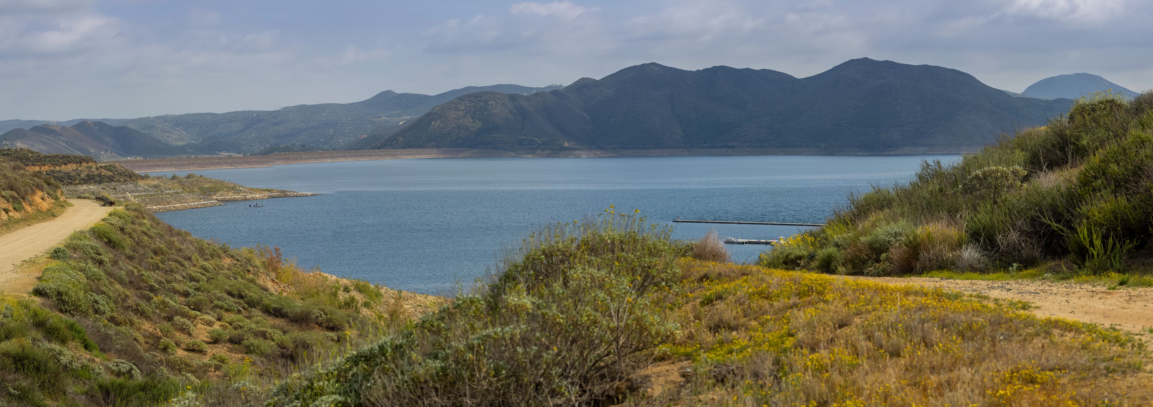 A coastal landscape with a dirt path, a calm lake, and mountains in the background.
