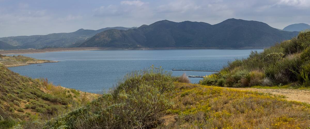 A coastal landscape with a dirt path, a calm lake, and mountains in the background.
