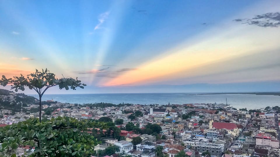 A coastal cityscape with a tree in the foreground and a sunset over the sea.