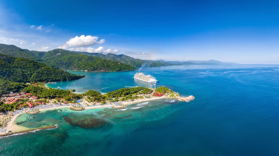 A cruise ship docked at a tropical island with clear turquoise waters, lush green hills, and a beach with umbrellas.