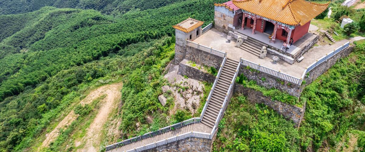 A traditional Chinese building with a tiled roof, situated on a hillside with lush greenery and a winding road in the background.