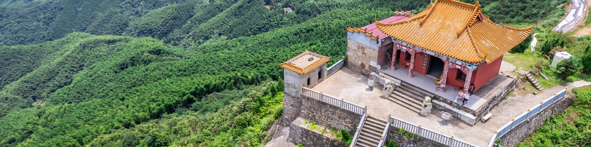 A traditional Chinese building with a tiled roof, situated on a hillside with lush greenery and a winding road in the background.