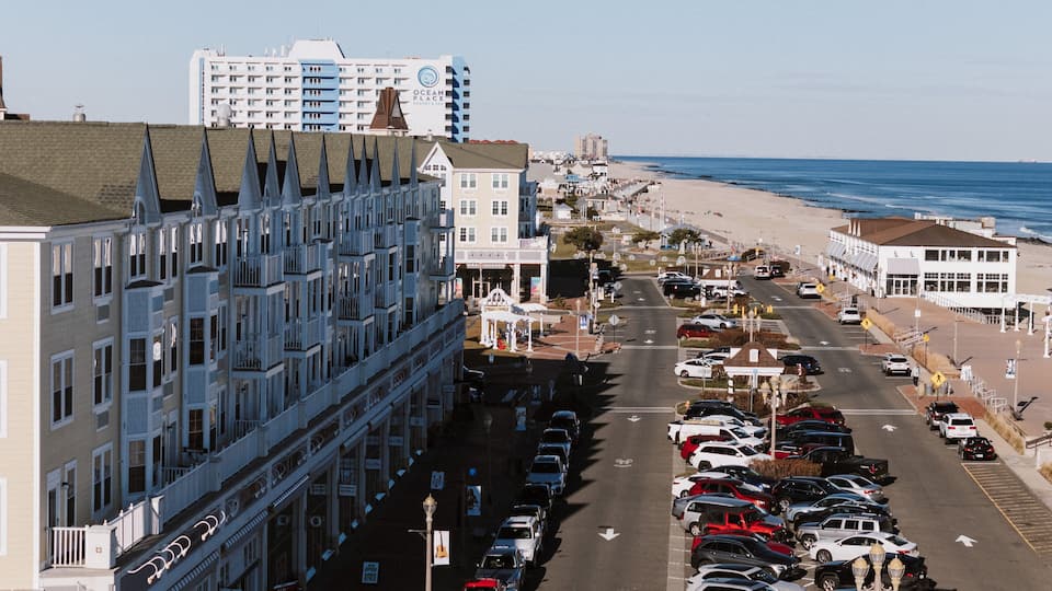 A coastal street lined with buildings, a beach, and parked cars.