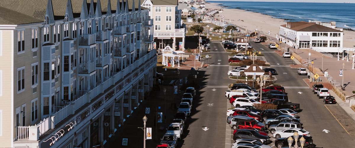 A coastal street lined with buildings, a beach, and parked cars.