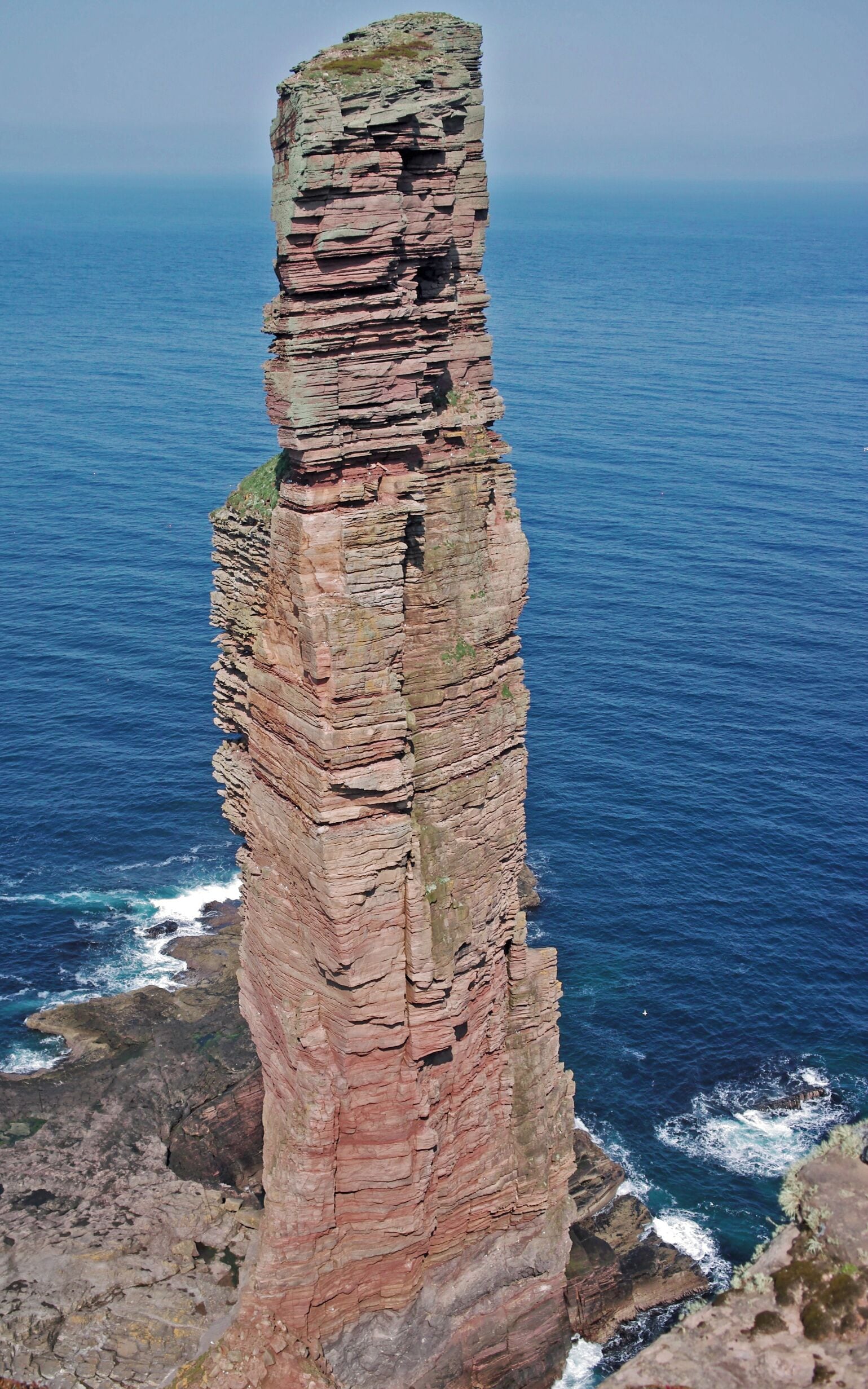 A tall, narrow rock formation with layered red and gray strata, overlooking a deep blue ocean.