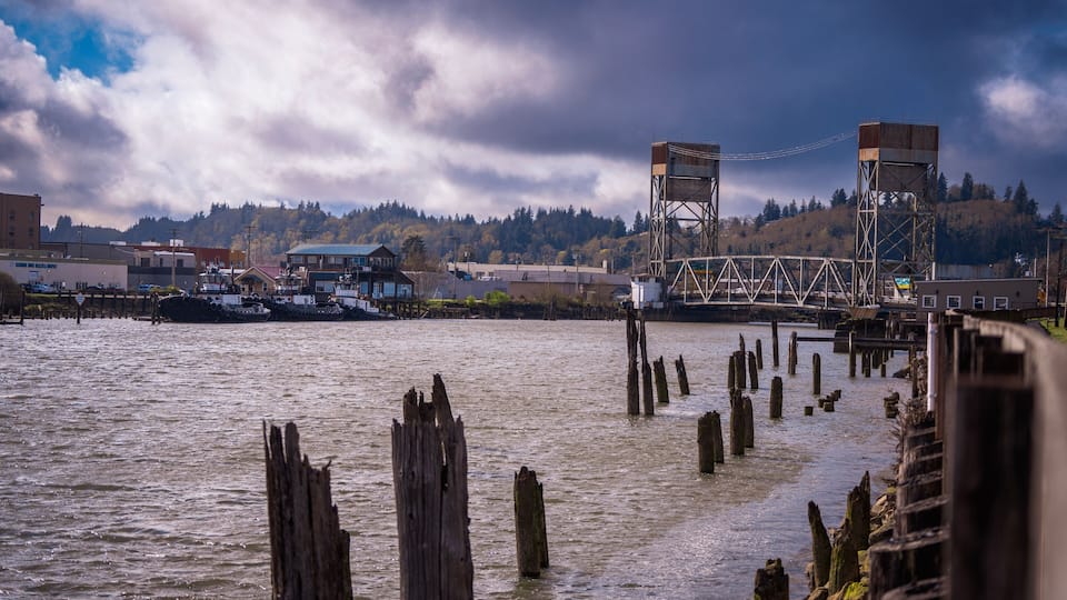 A river with wooden posts, a bridge, and buildings in the background.