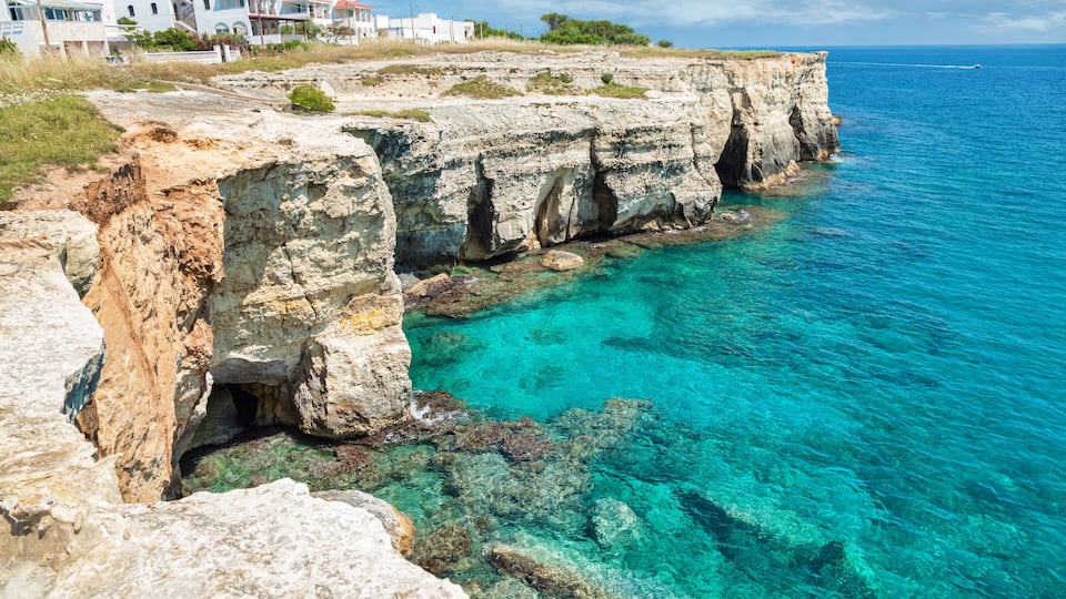 A coastal cliff with clear turquoise water, rocky formations, and houses in the background.