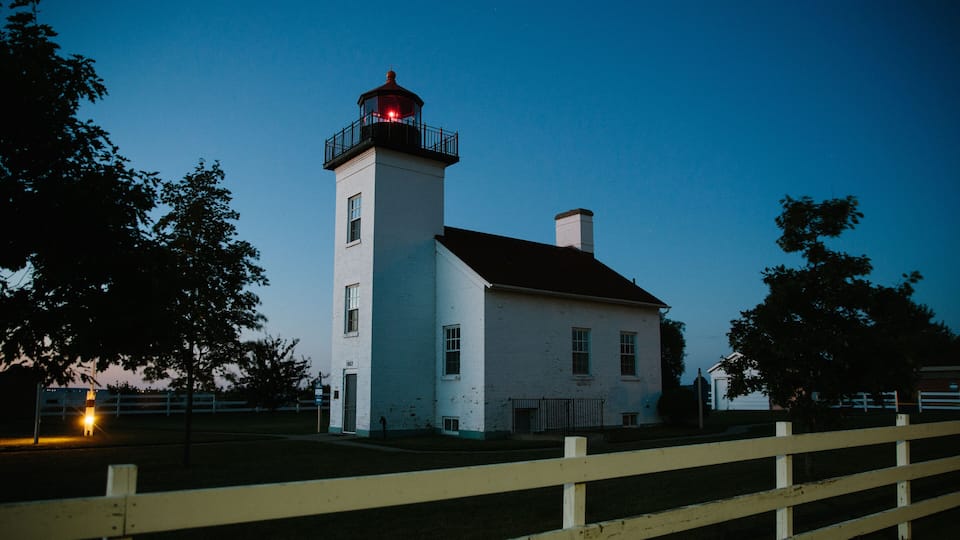 A white lighthouse with a red light atop, surrounded by trees and a wooden fence.