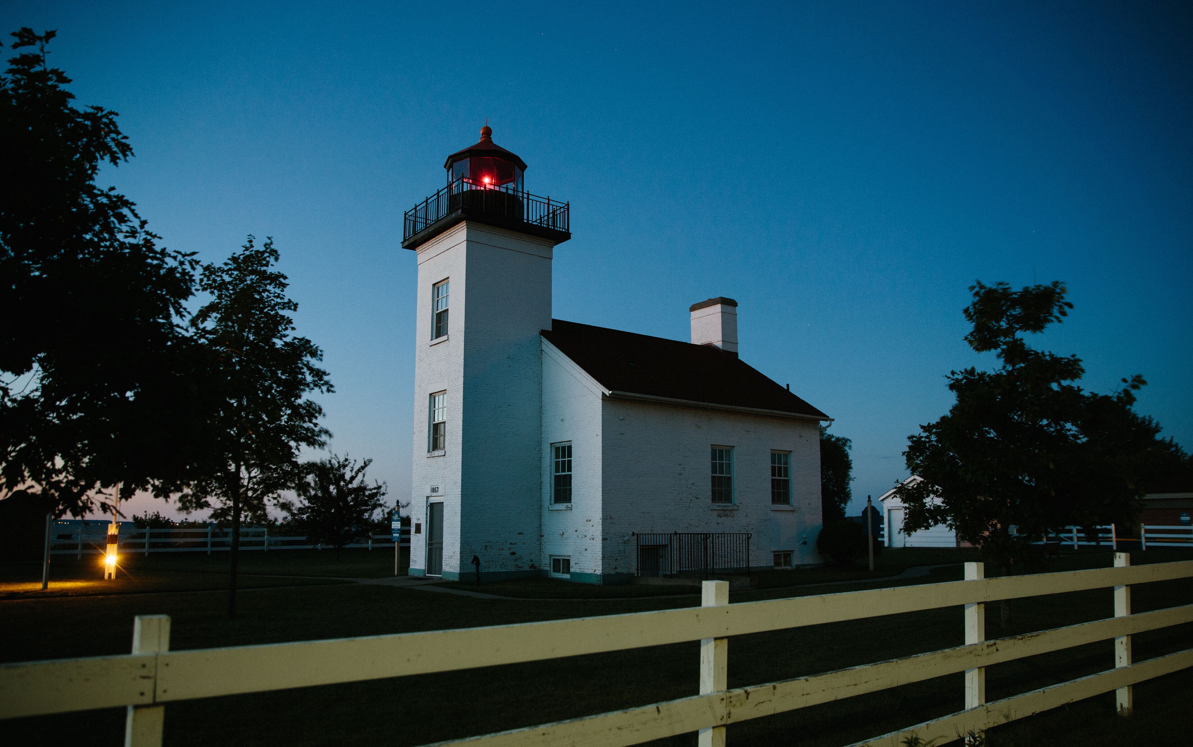 A white lighthouse with a red light atop, surrounded by trees and a wooden fence.
