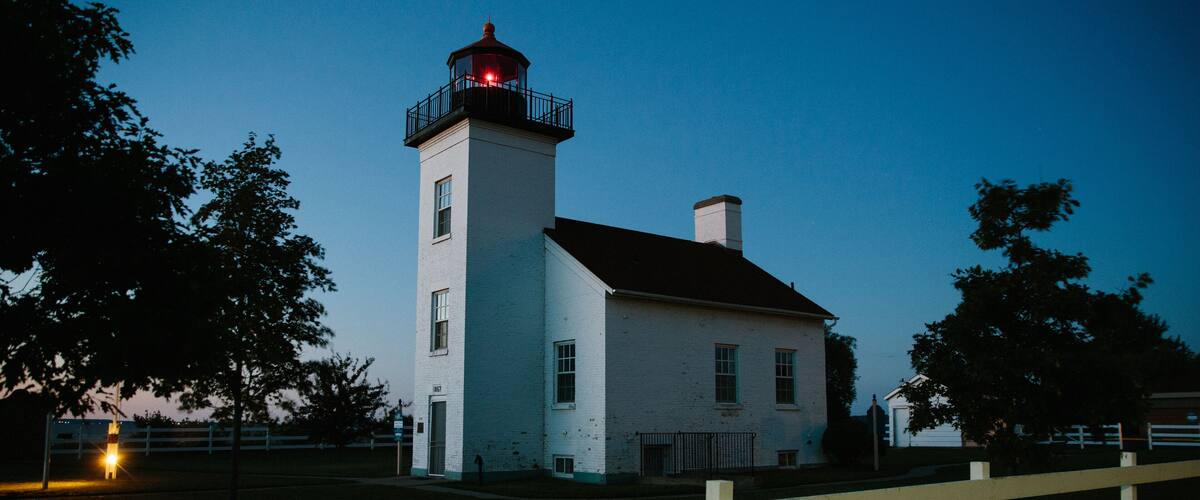 A white lighthouse with a red light atop, surrounded by trees and a wooden fence.