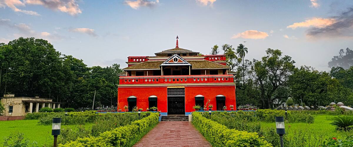 A red building with a tiled roof and a central entrance, surrounded by green hedges and trees.